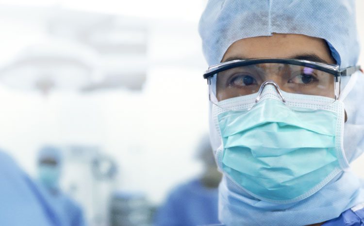 Closeup photo of a surgeon's face in scrubs, protective glasses and mask, looking away from camera.