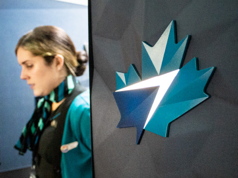 Image of the cabin interior of a WestJet's airplane. A lit-up WestJet maple leaf logo can be seen on the wall, and a flight attendant is passing by in the background.